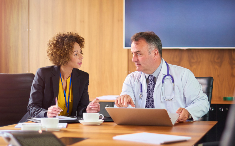 a senior doctor chats to a woman in business wear at a conference table. They are discussing something on their laptop . She could be the hospital administrator , salesperson or lawyer .