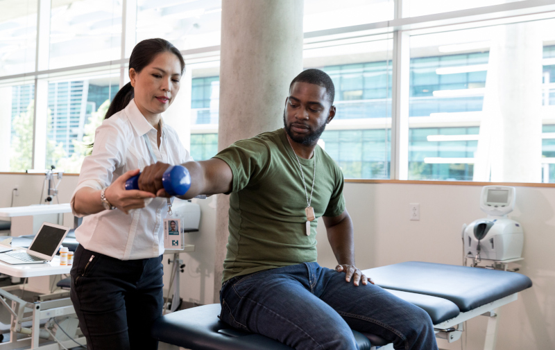 Serious army soldier with injury uses a hand weight during a physical therapy session. A female physical therapist is helping him with an exercise.